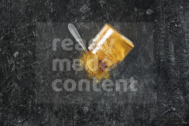 A flipped glass jar and a metal spoon full of turmeric powder and powder spilled out of it on textured black flooring