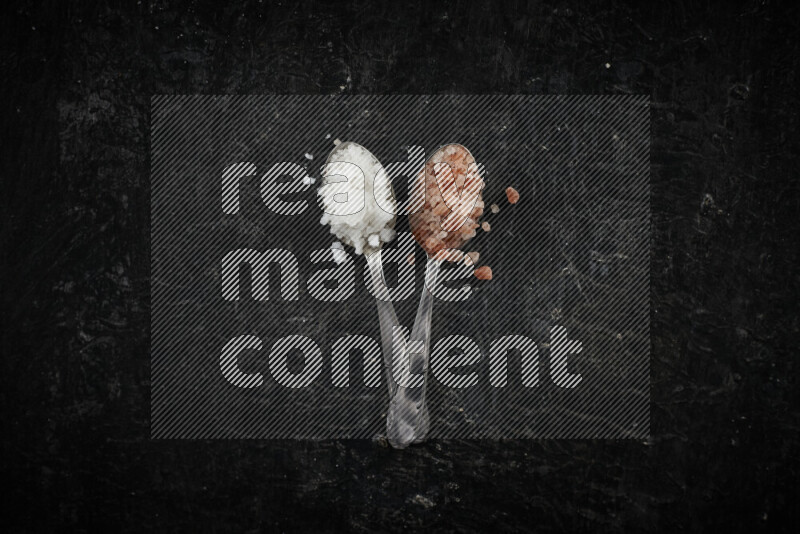 ملعقتان معدنيتان ممتلئتان بالملح الأبيض وملح الهيمالايا الوردي على خلفية سوداء