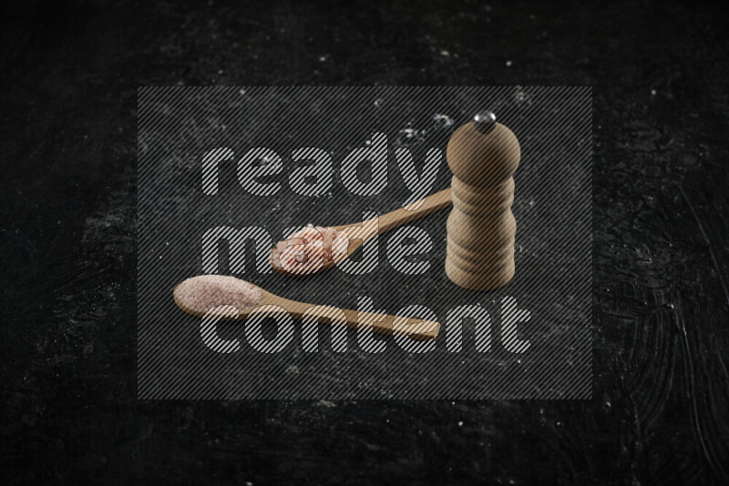2 wooden spoons filled with fine and coarse salt with wooden grinder beside them on black background