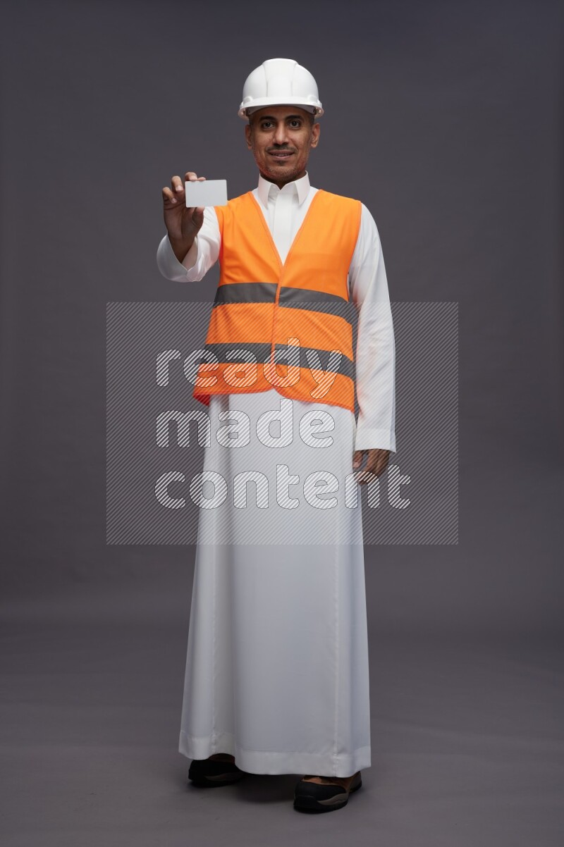 Saudi man wearing thob with engineer vest standing holding ATM card on gray background