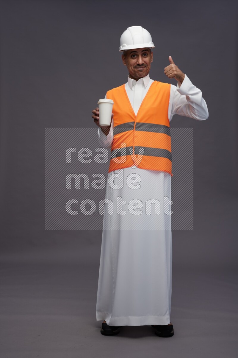 Saudi man wearing thob with engineer vest standing holding paper cup on gray background