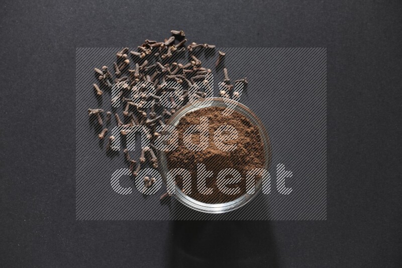 A glass bowl full of cloves powder with cloves grains spread on black flooring