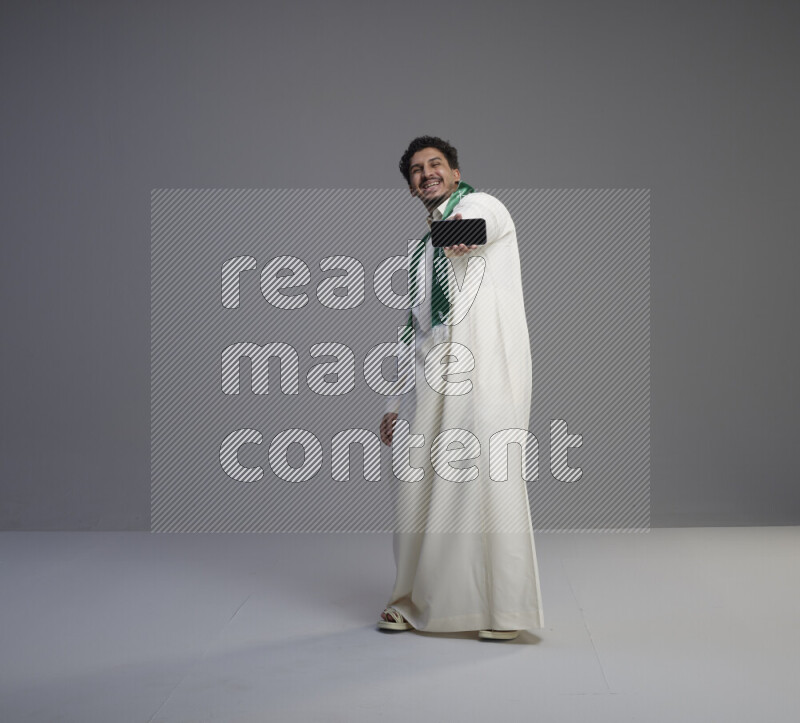 A Saudi man standing wearing thob and Saudi flag scarf showing phone to camera on gray background