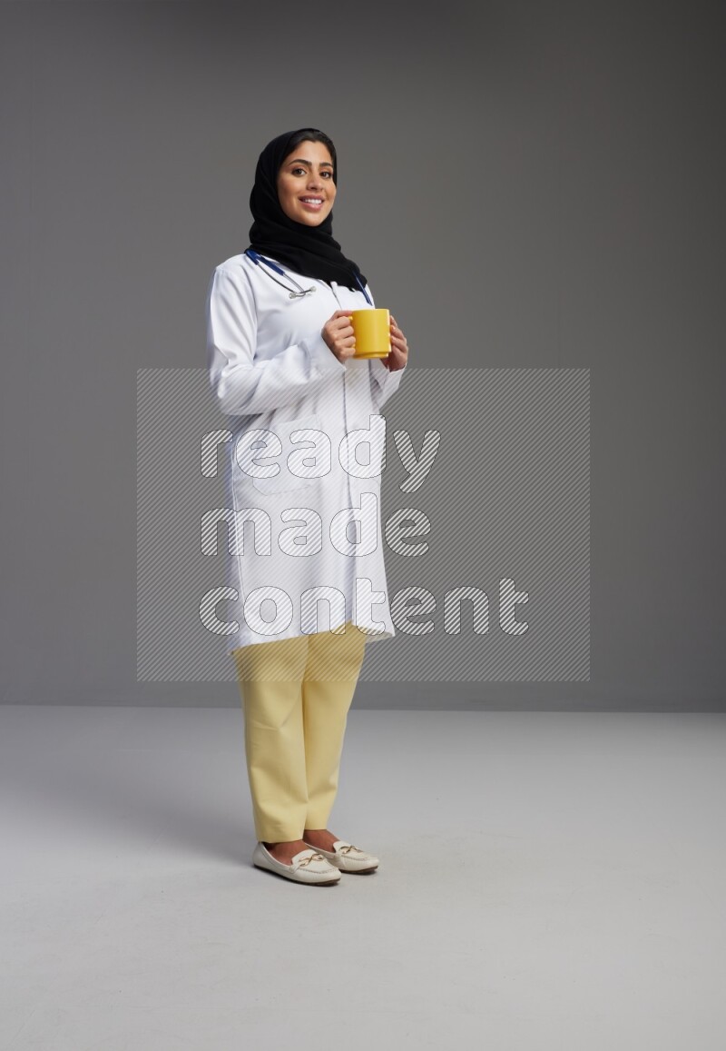 Saudi woman wearing lab coat with stethoscope standing holding mug on Gray background