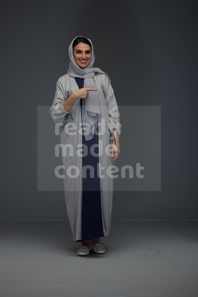 A Saudi woman wearing a light gray Abaya and head scarf standing and pointing in different directions eye level on a grey background