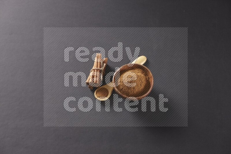 Cinnamon sticks stacked and bounded beside a wooden bowl full of cinnamon powder and a wooden spoon full of powder on black background