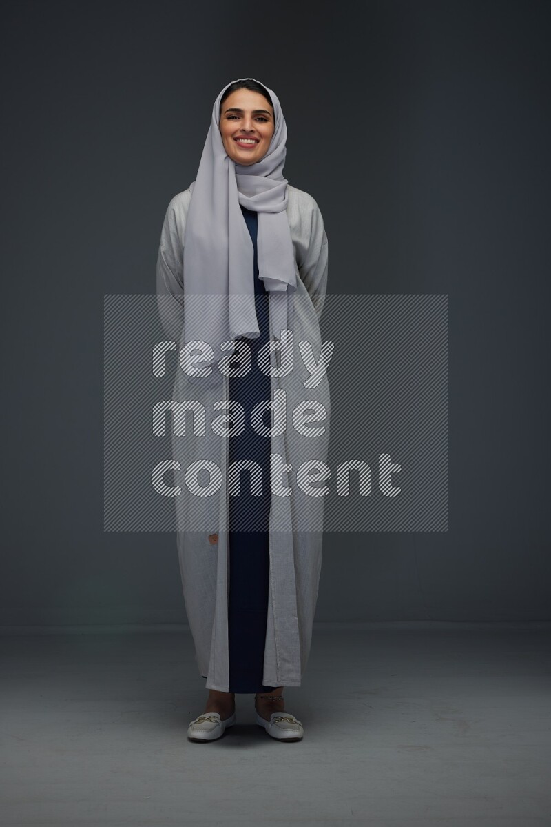 A Saudi woman wearing a light gray Abaya and head scarf standing and making multi poses eye level on a grey background