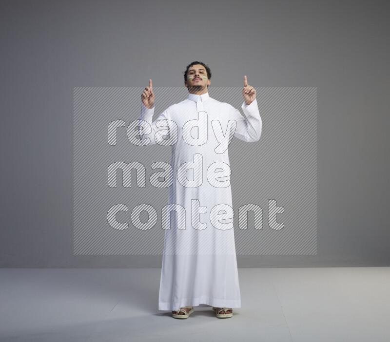 A Saudi man standing wearing thob with face painting interacting with the camera on gray background