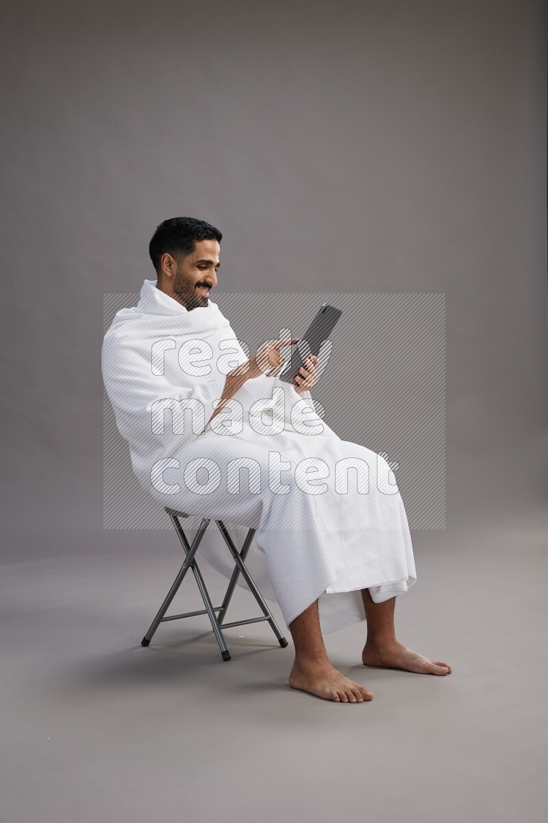 A man wearing Ehram sitting on chair holding a tablet on gray background