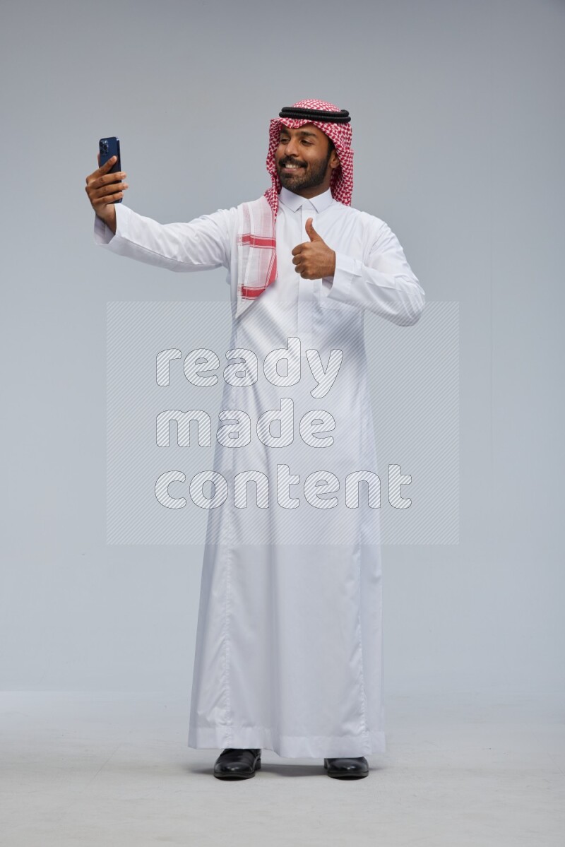 Saudi man Wearing Thob and shomag standing taking selfie on Gray background