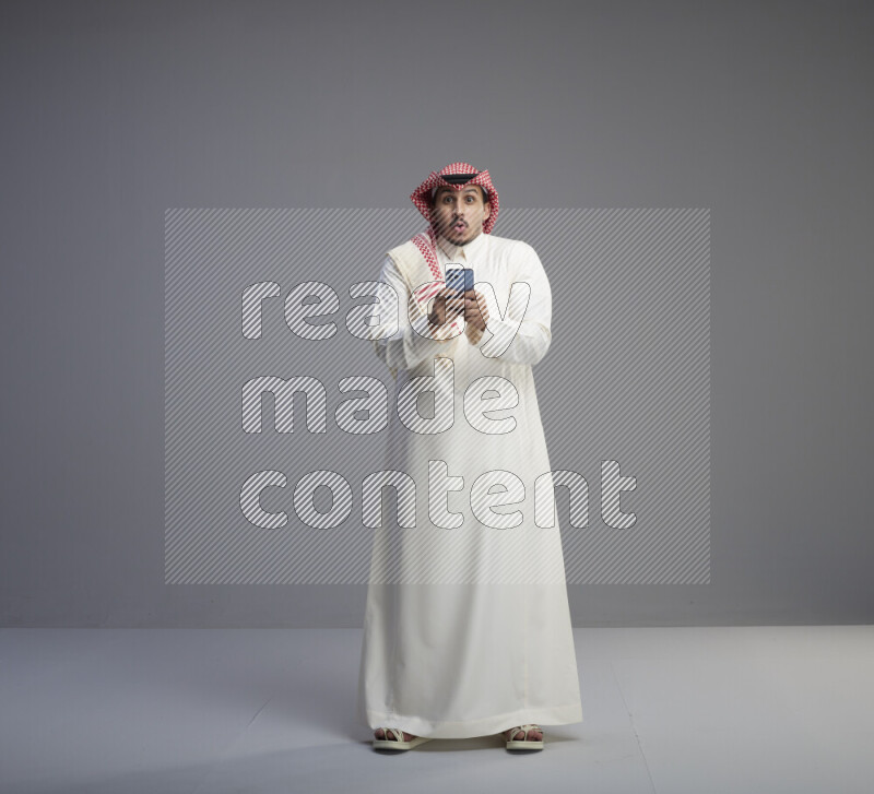 A  Saudi man standing wearing thob and red shomag texting on phone on gray background