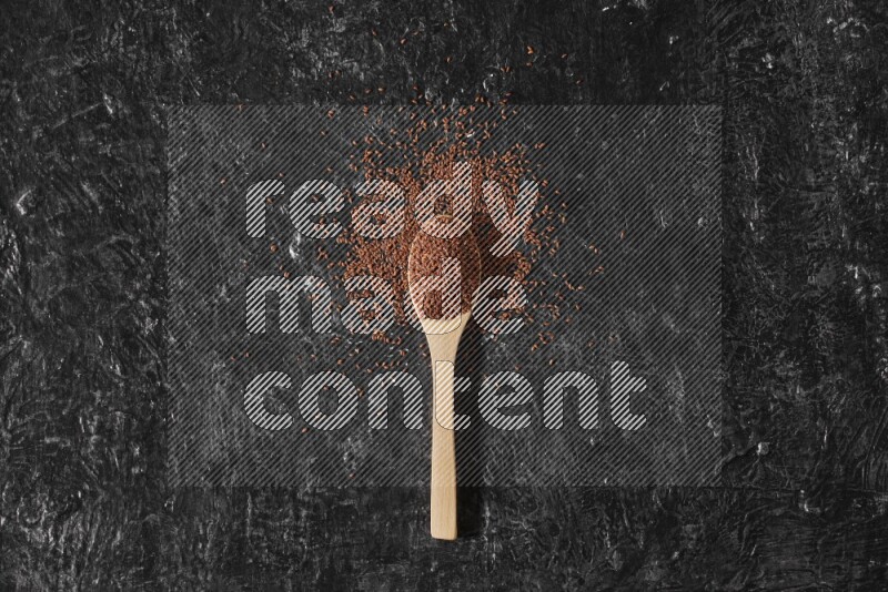 A wooden spoon full of garden cress seeds surrounded by the seeds on a textured black flooring