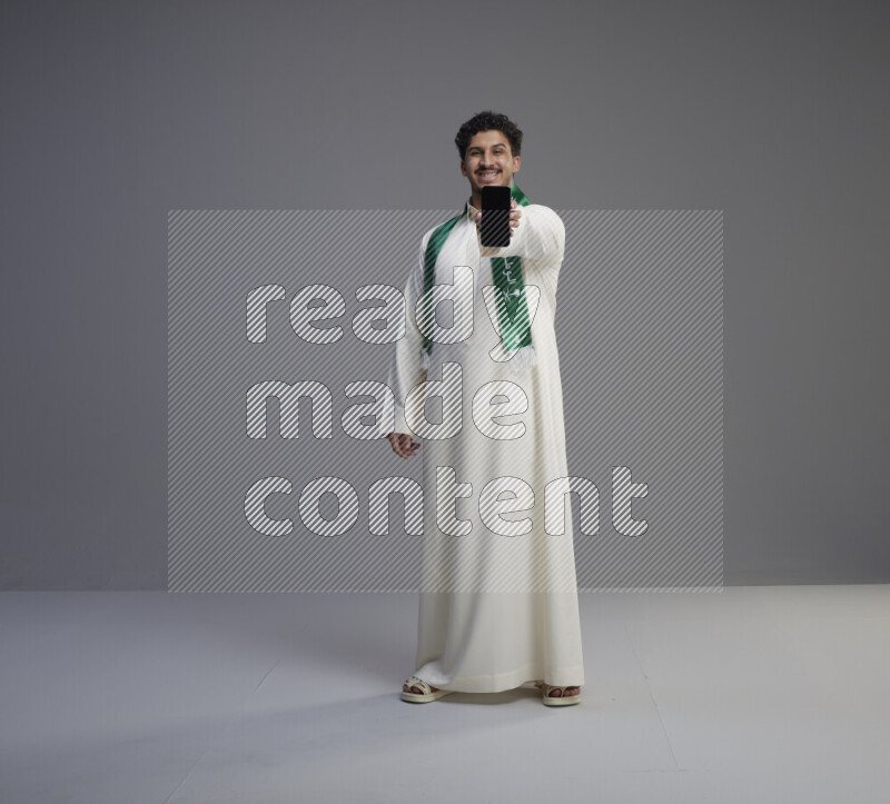 A Saudi man standing wearing thob and Saudi flag scarf showing phone to camera on gray background