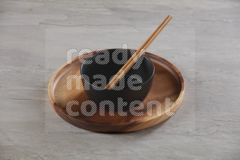 black ceramic round bowl on top of brown wood round plate and wood chopsticks, on grey textured countertop