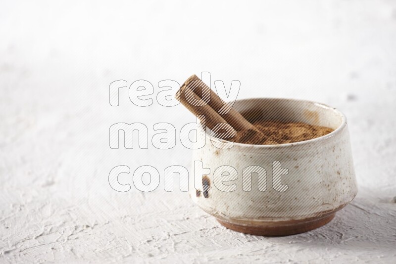 Ceramic beige bowl full of cinnamon powder with a cinnamon stick on a textured white background