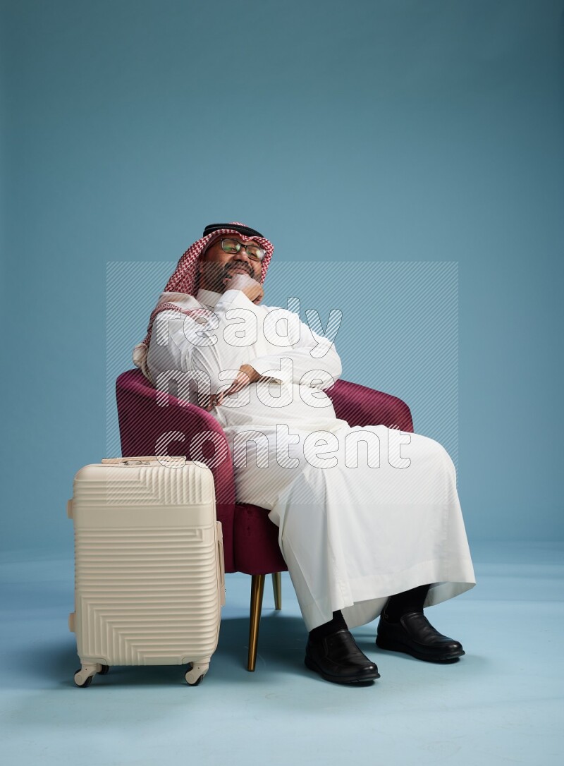 Saudi Man with shimag sitting on chair with a travel luggage on blue background