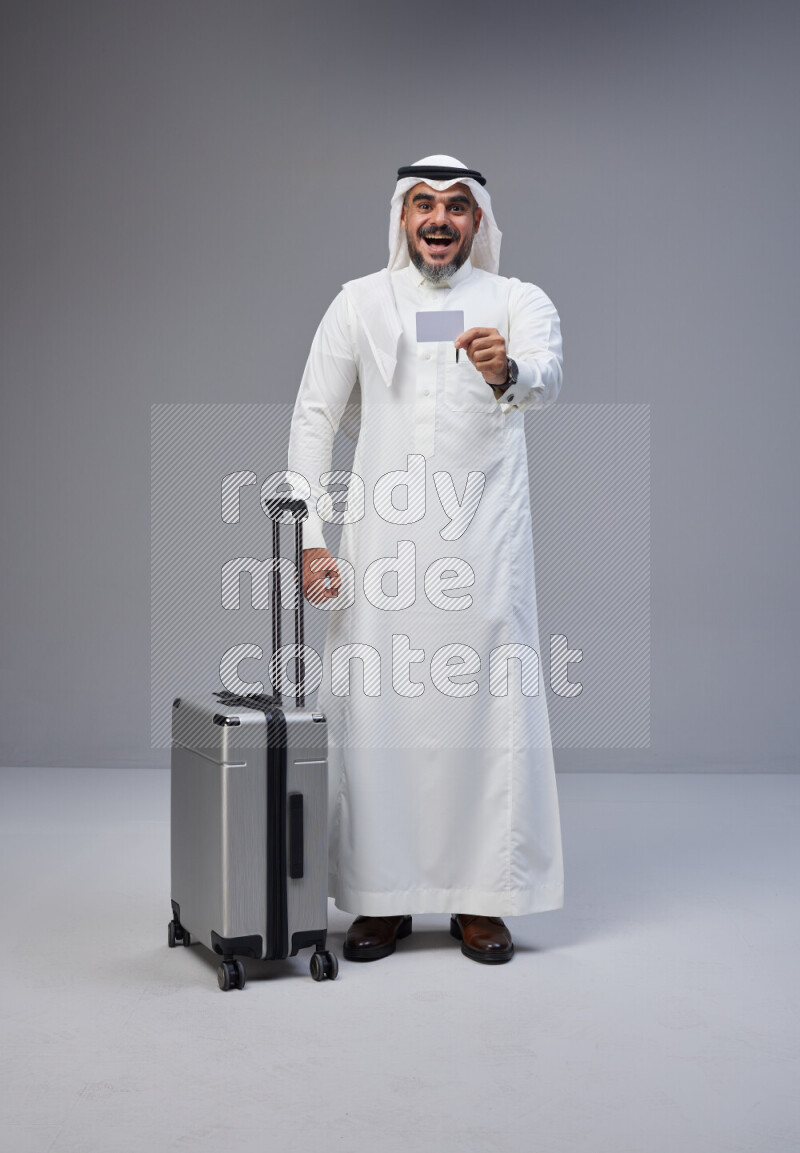 Saudi man wearing Thob and white Shomag standing holding Travel bag and ATM card on Gray background