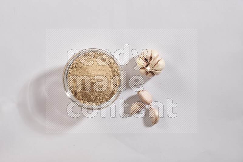 A glass bowl full of garlic powder with garlic bulb and some cloves beside it on a white flooring