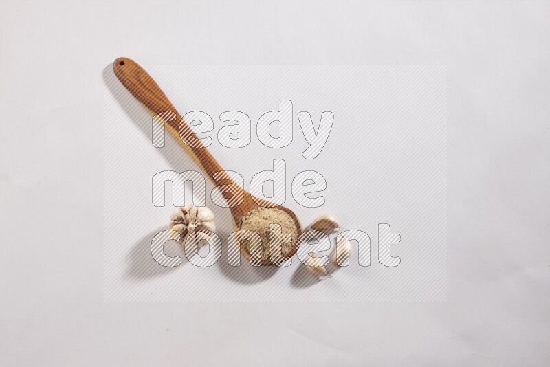 A wooden ladle full of garlic powder with a whole garlic bulb and some cloves beside it on a white flooring