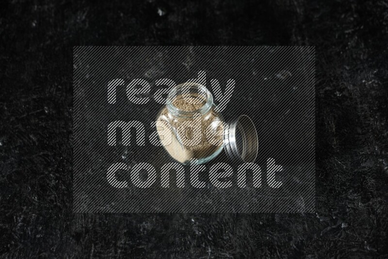 A glass spice jar full of cardamom powder on textured black flooring