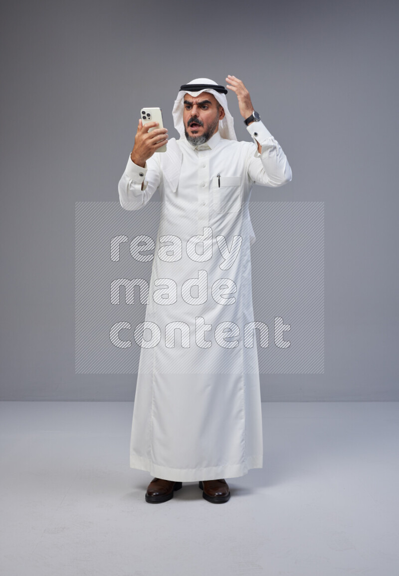 Saudi man Wearing Thob and white Shomag standing texting on phone on Gray background