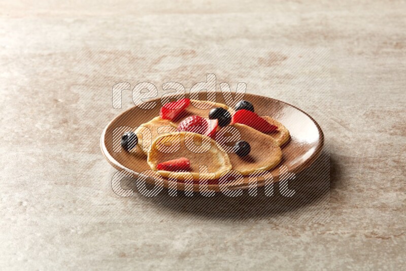 Five stacked mixed berries mini pancakes in a brown plate on beige background