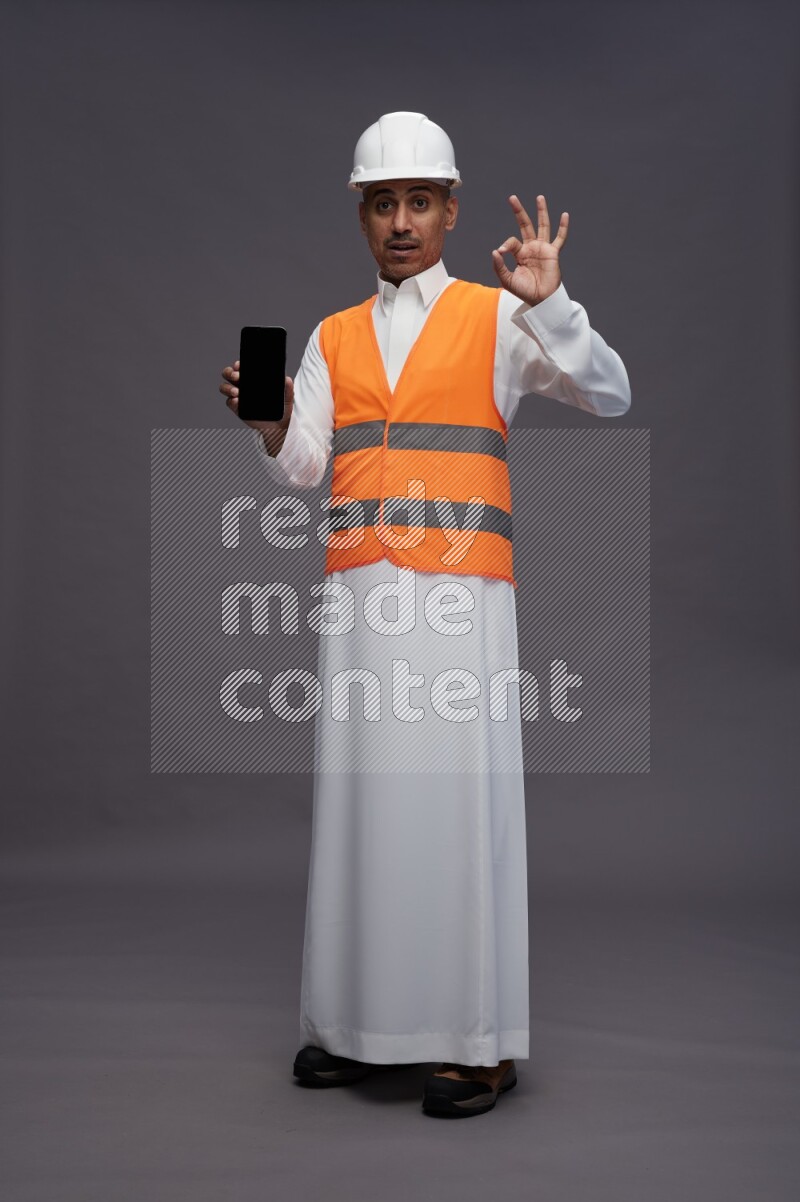Saudi man wearing thob with engineer vest standing showing phone to camera on gray background