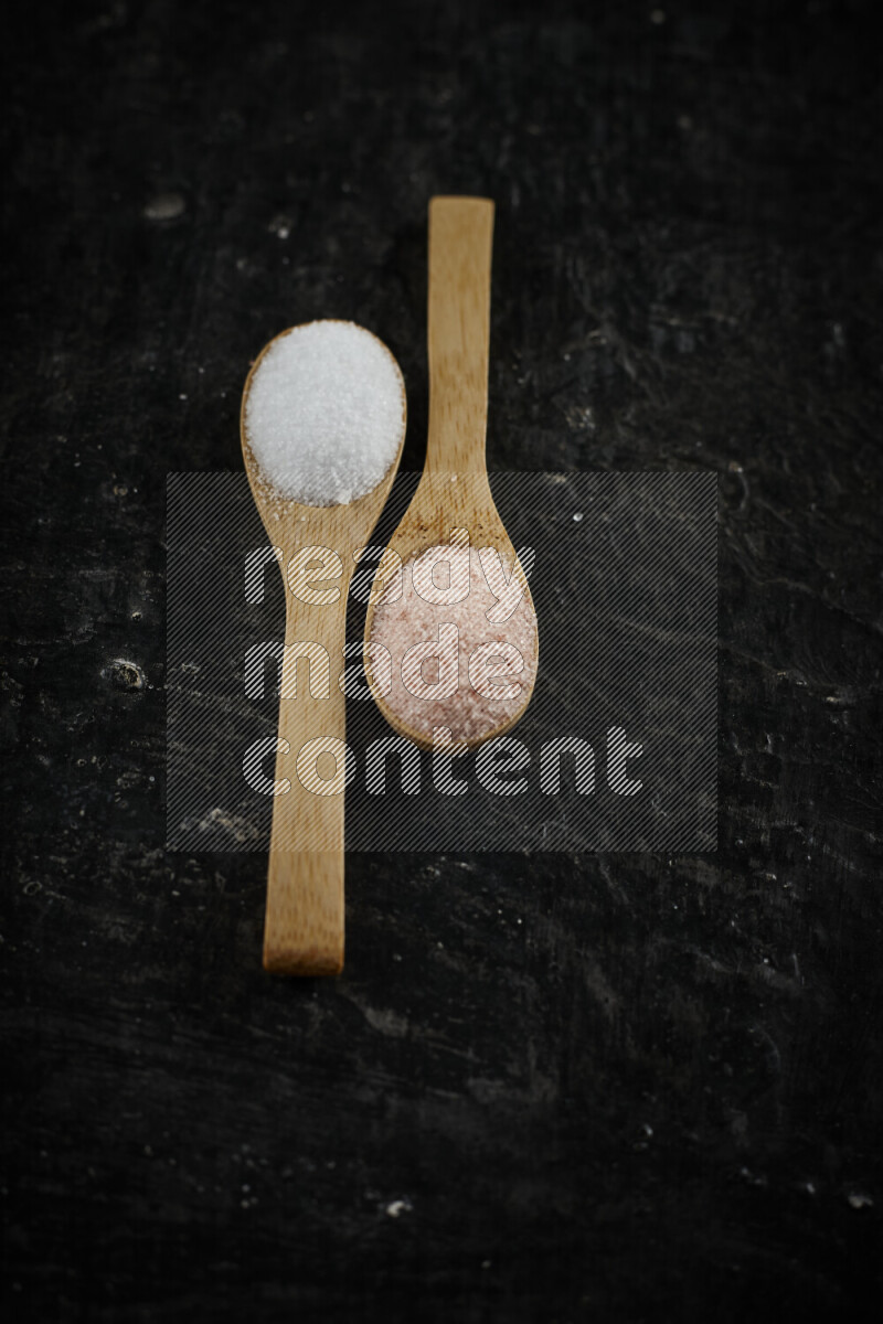 2 wooden spoons filled with fine and coarse salt on black background