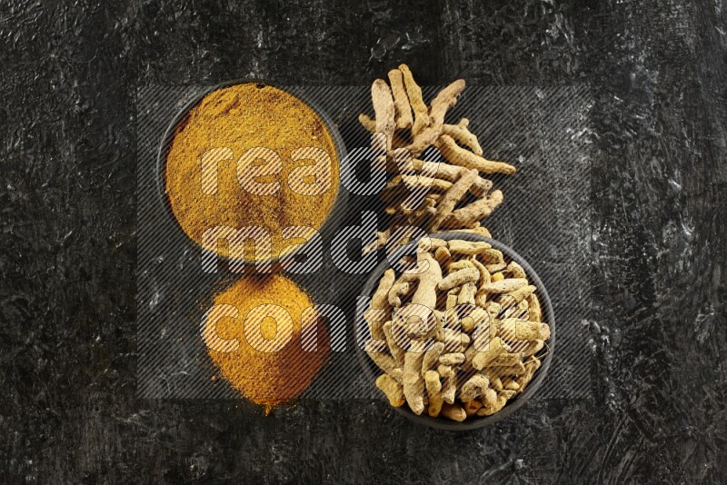 2 pottery black bowls, one full of turmeric powder and the other full of dried turmeric whole fingers on a textured black flooring
