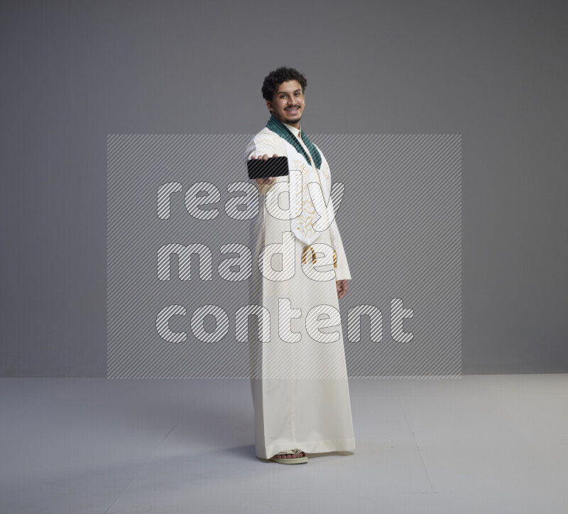 A Saudi man standing wearing thob and Saudi flag scarf showing phone to camera on gray background