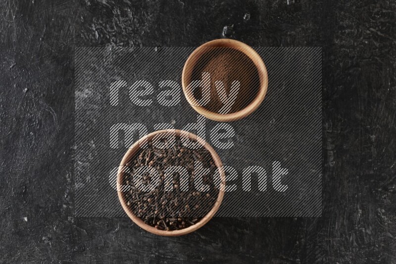 2 wooden bowls full of cloves powder and whole cloves on a textured black flooring