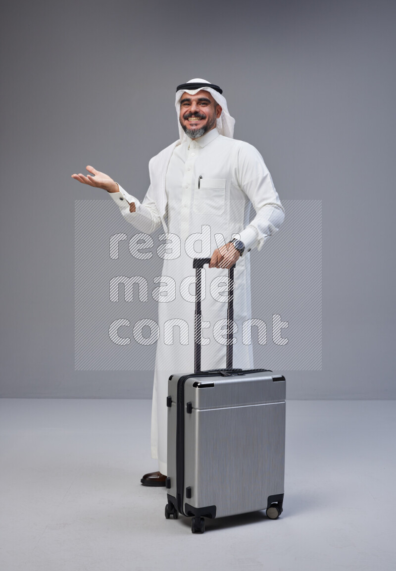 Saudi man wearing Thob and white Shomag standing holding Travel bag on Gray background