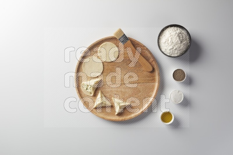 two closed sambosas and one open sambosa filled with cheese while flour, salt, black pepper and oil with oil brush aside in a wooden dish on a white background