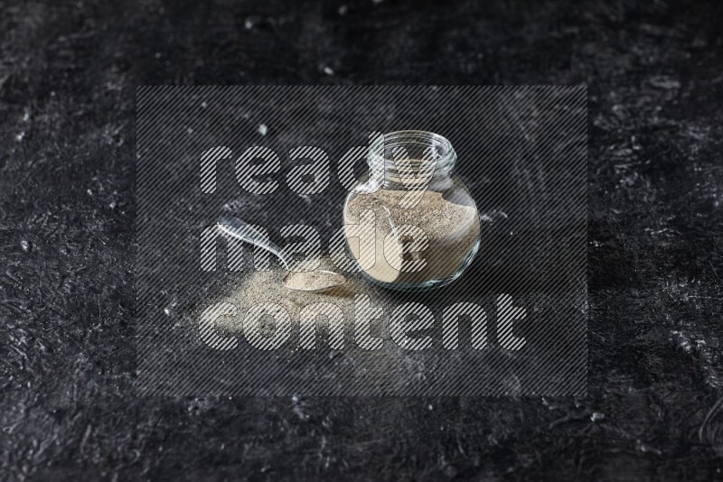 Herbal glass jar and metal spoon full of white pepper powder on textured black flooring