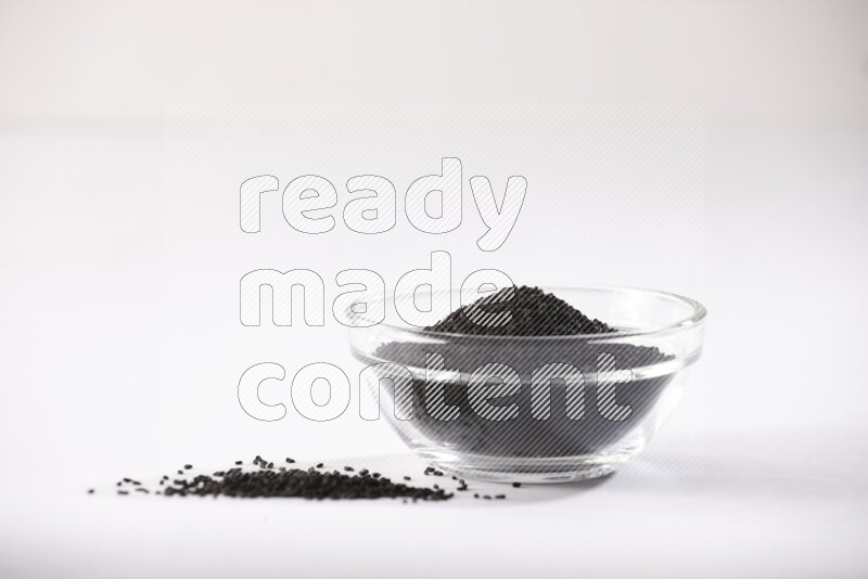 A glass bowl full of black seeds and some more seeds spread next to it on a textured white flooring
