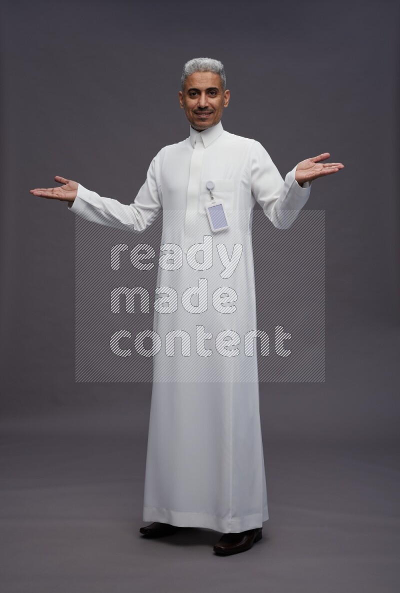 Saudi man wearing thob with pocket employee badge standing interacting with the camera on gray background