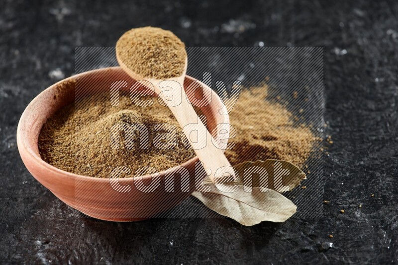 A wooden bowl and spoon full of cumin powder on a textured black flooring