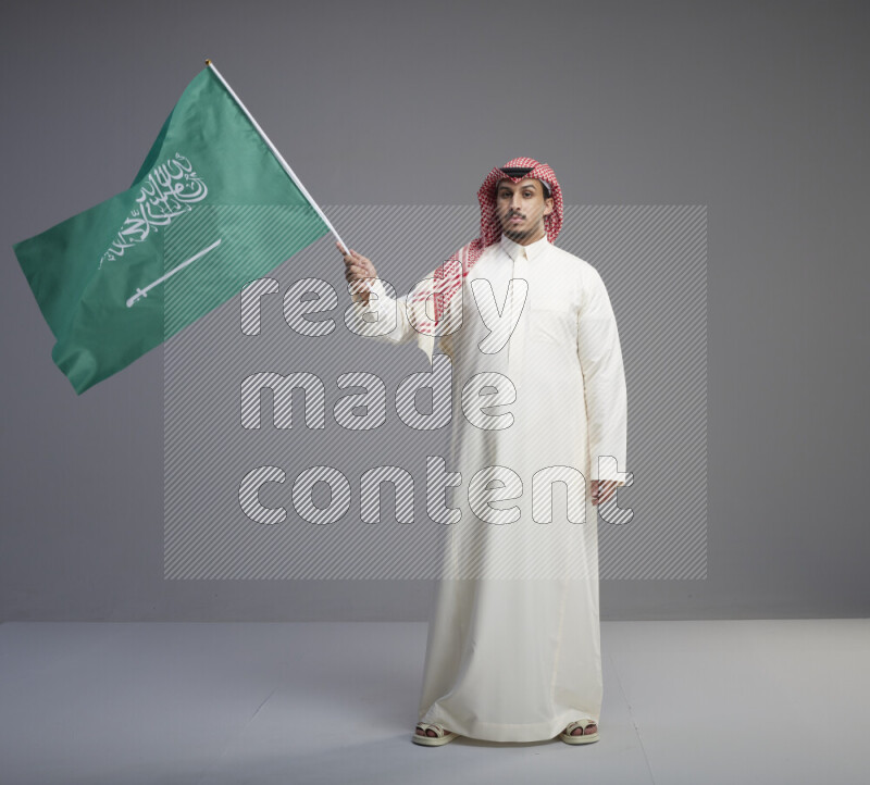 A Saudi man standing wearing thob and red shomag raising big Saudi flag on gray background