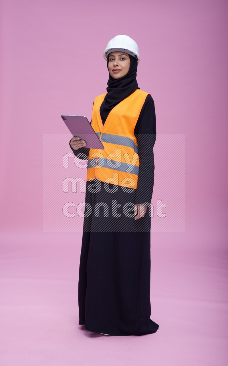 Saudi woman wearing Abaya with engineer vest and helmet standing working on tablet on pink background