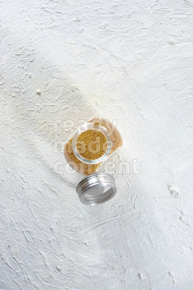 A glass spice jar full of turmeric powder on a textured white flooring