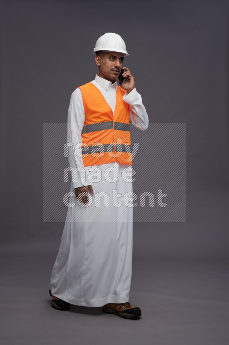 Saudi man wearing thob with engineer vest standing talking on phone on gray background