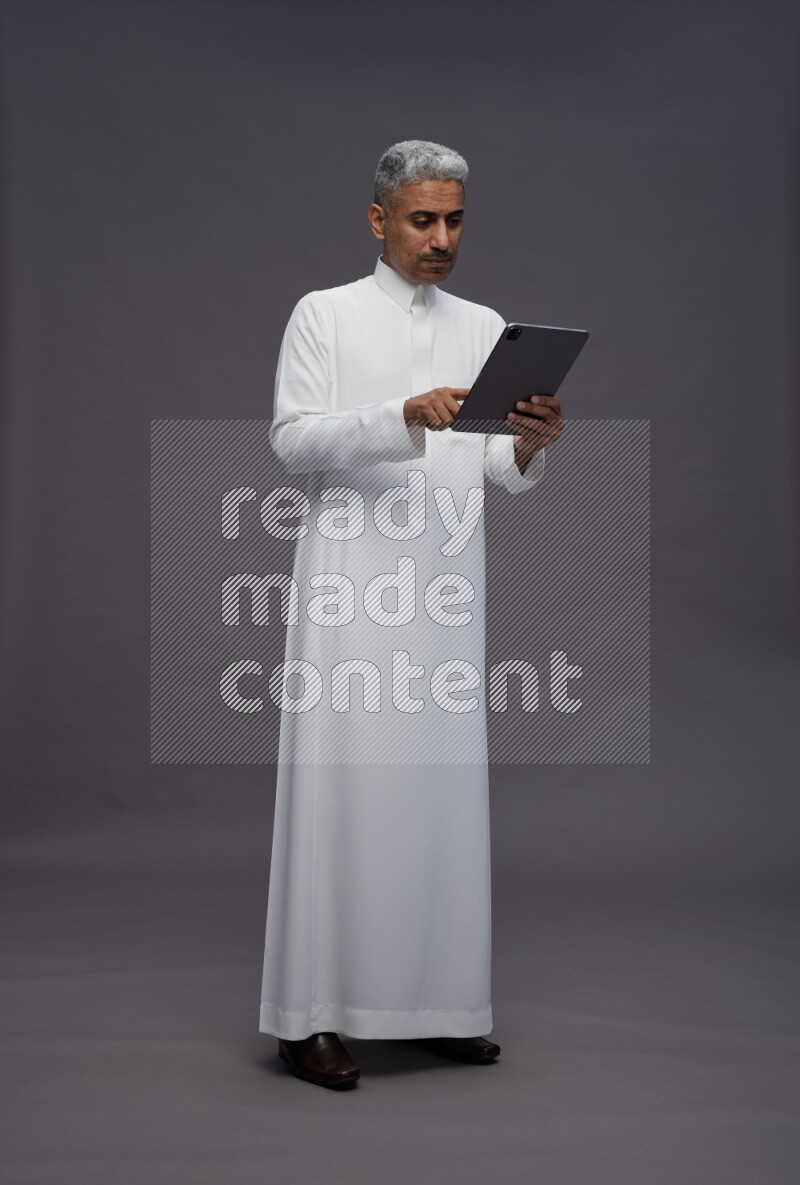 Saudi man wearing thob standing working on tablet on gray background