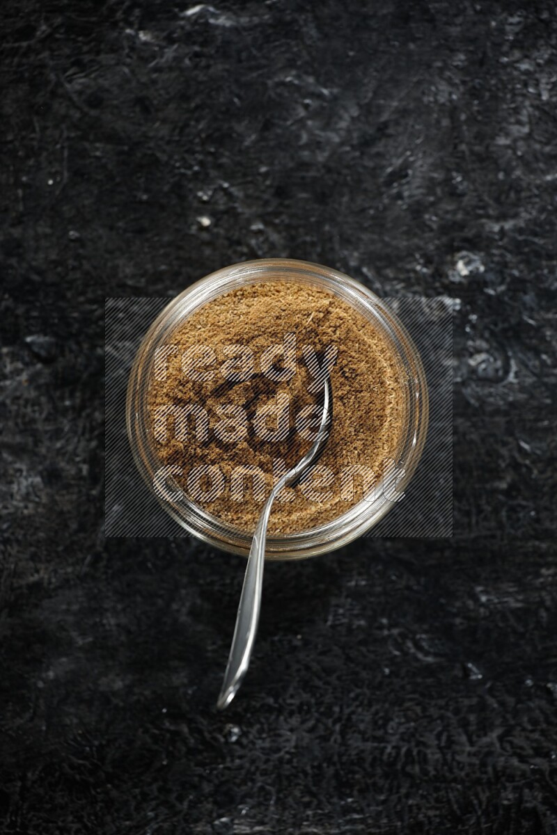 A glass jar and a metal spoon full of cumin powder on a textured black flooring