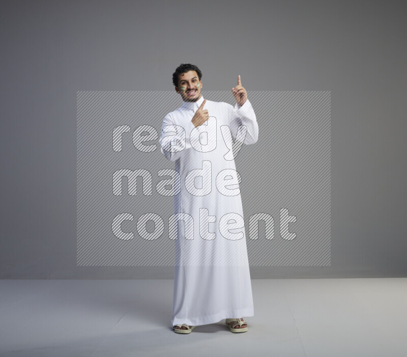 A Saudi man standing wearing thob with face painting interacting with the camera on gray background