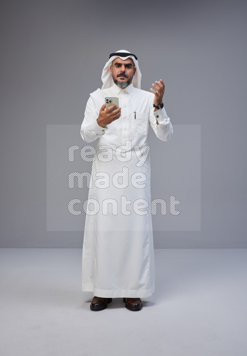 Saudi man Wearing Thob and white Shomag standing texting on phone on Gray background