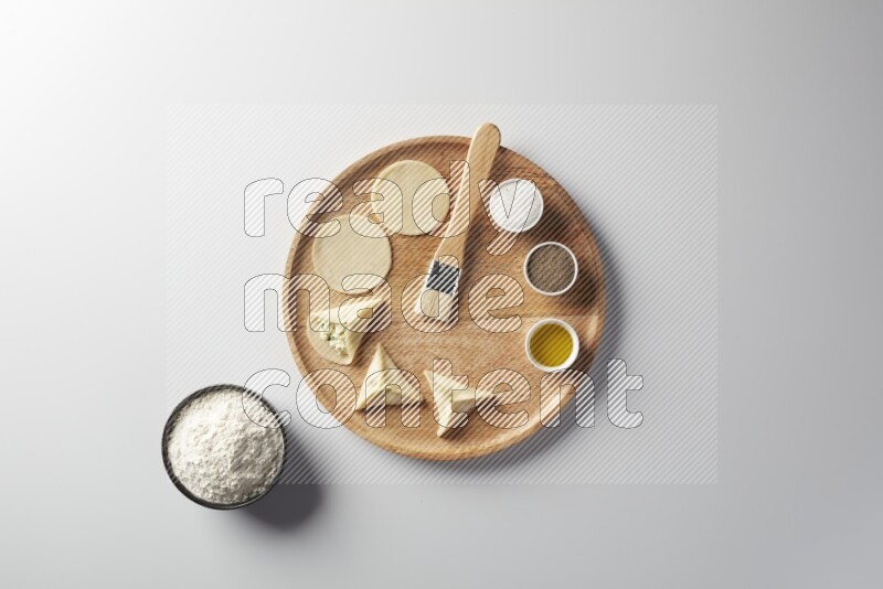 two closed sambosas and one open sambosa filled with cheese while flour, salt, black pepper and oil with oil brush aside in a wooden dish on a white background