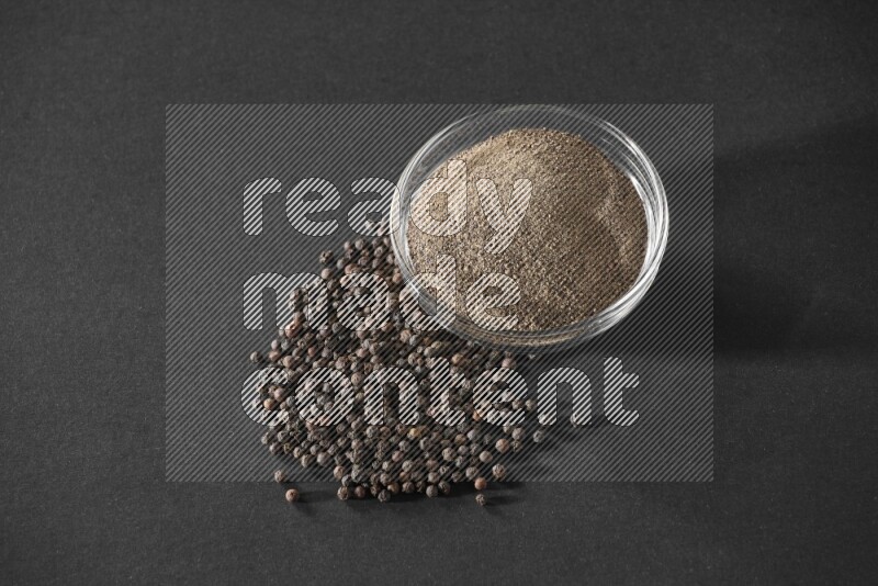 A glass bowl full of powder black pepper powder and black pepper beads on the floor on black flooring