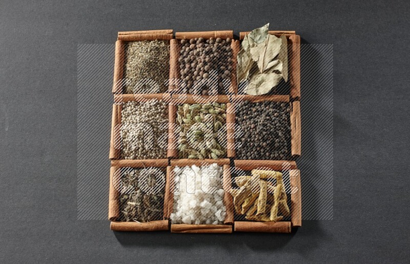 9 squares of cinnamon sticks full of cardamom in the middle surrounded by turmeric fingers, black pepper, bay laurel leaves, allspice, cumin, white pepper, dried basil and salt on black flooring