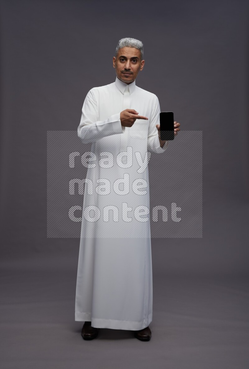 Saudi man wearing thob standing showing phone to camera on gray background