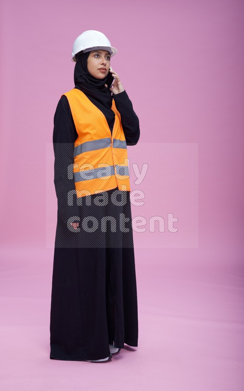 Saudi woman wearing Abaya with engineer vest and helmet standing talking on phone on pink background